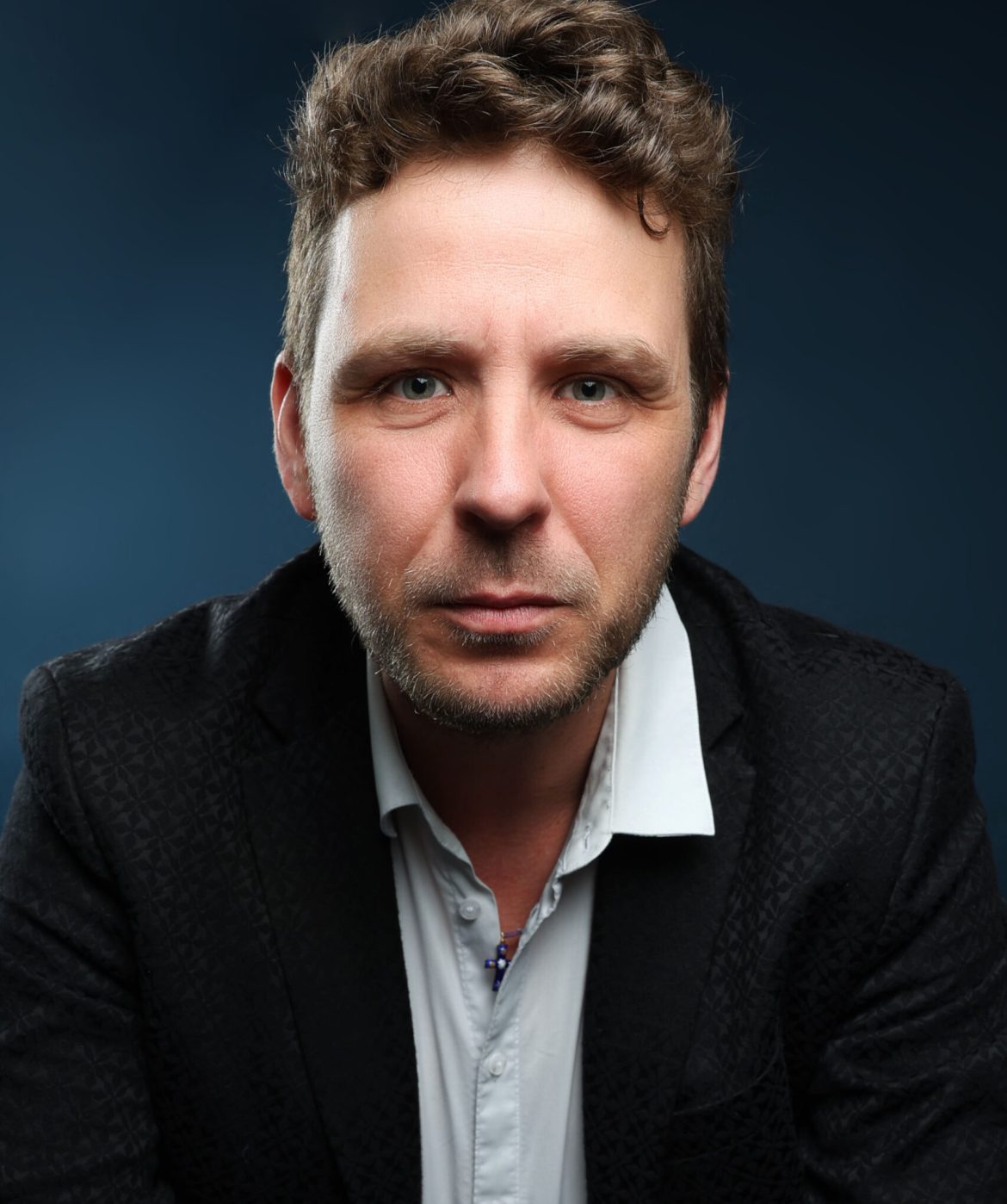 Portrait professionnel d’un thérapeute bioénergéticien, veste noire et chemise blanche, fond bleu studio