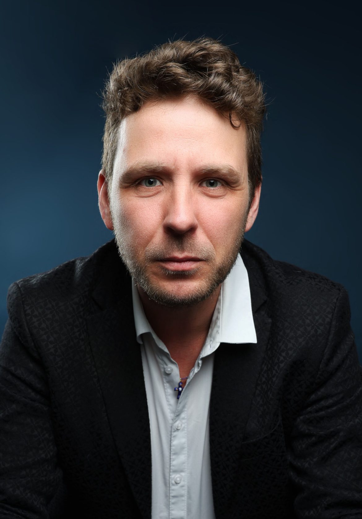 Portrait professionnel d’un thérapeute bioénergéticien, veste noire et chemise blanche, fond bleu studio